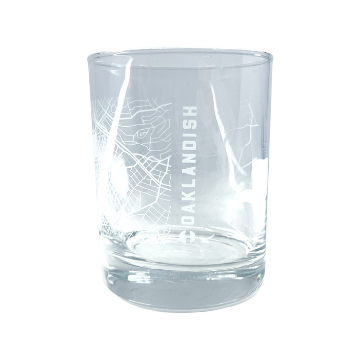 The Aerial Tumbler by Oaklandish is a clear glass whiskey tumbler with a minimalist design that boasts an etched, abstract aerial map of Oakland. The word "OAKLANDISH" is prominently displayed in vertical capital letters down one side of the classic cylindrical glass, which features a thick base.