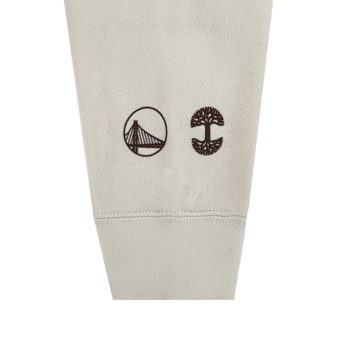 All Gold Forever Crew by Other features a beige fabric sleeve adorned with two minimalist black icons near the cuff: a suspension bridge inside a circle on the left and a stylized tree with an expansive canopy and visible roots on the right. This crewneck sweatshirt is showcased against a white background.