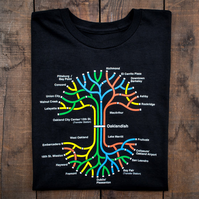 The Oaklandish BART Tree Tee, made from 100% black cotton, is displayed on a wooden surface. This classic fit t-shirt features a colorful, stylized BART map of the San Francisco Bay Area with stations like Civic Center, 12th St., and Concord. The design showcases lines in red, yellow, blue, green, and orange.