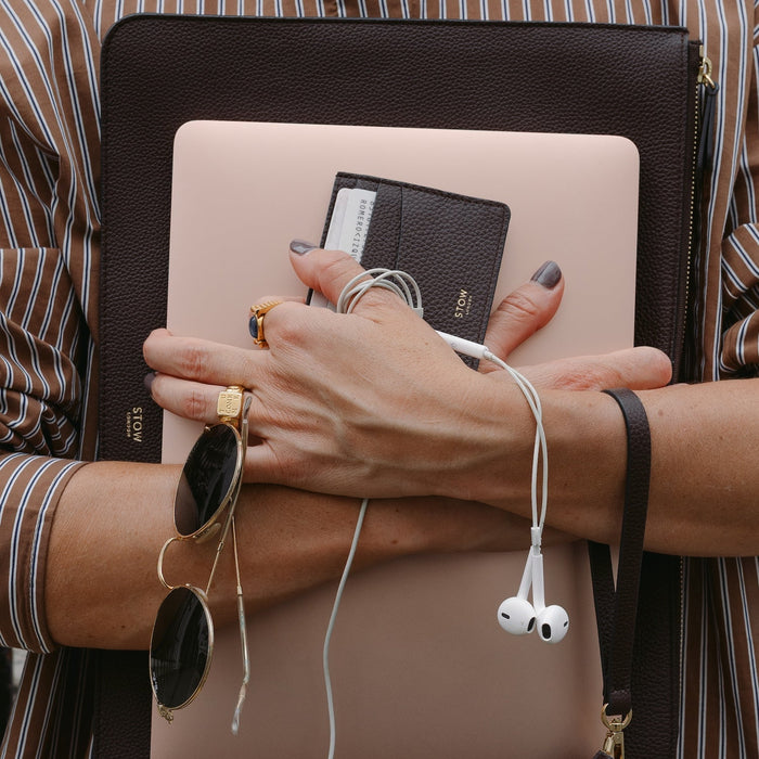 Woman holding STOW London's Leather Sleeve and Cardholder in Espresso pebbled leather, alongside a peach laptop, sunglasses and earphones.