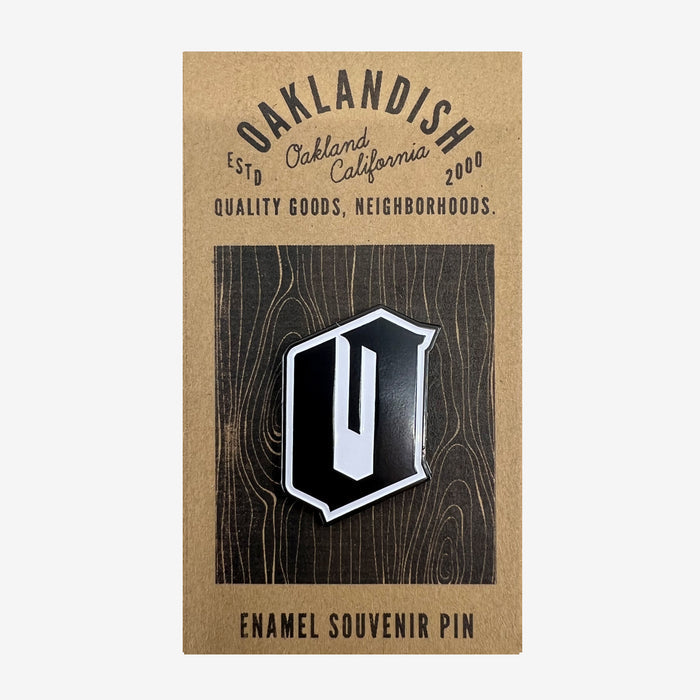 A black enamel lapel pin featuring a silver-bordered, stylized letter "O" displayed on a wooden background card. The card reads "Oaklandish, Estd Oakland California 2000, Quality Goods, Neighborhoods," and ends with "Enamel Souvenir Accessories" at the bottom.