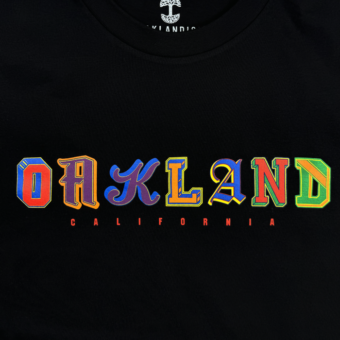The First Team Tee by Other is a black, heavy-weight cotton t-shirt featuring “OAKLAND” in large, colorful, uniquely designed letters across the chest. Beneath it, “CALIFORNIA” appears in small, spaced-out red letters. This classic fit tee has a rounded neckline for comfort and timeless style.