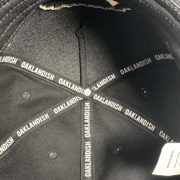 Close-up of the inside of a black Growth Cap by Oakland Roots SC. The inner seams feature black fabric strips displaying "OAKLANDISH" in white, and a white tag is partially visible along the rim under the light.