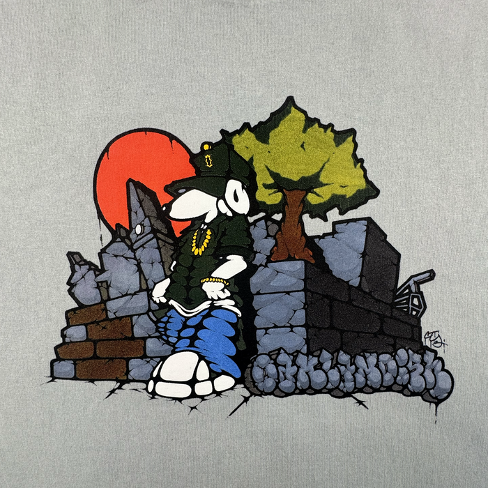 A cartoon dog in urban style leans by cracked ruins and a tree, wearing a green hat, necklace, blue jeans, and the Oaklandish Highroller Tee—a vintage grey classic fit. He stands relaxed as a large red sun sets in this street-art-inspired scene.