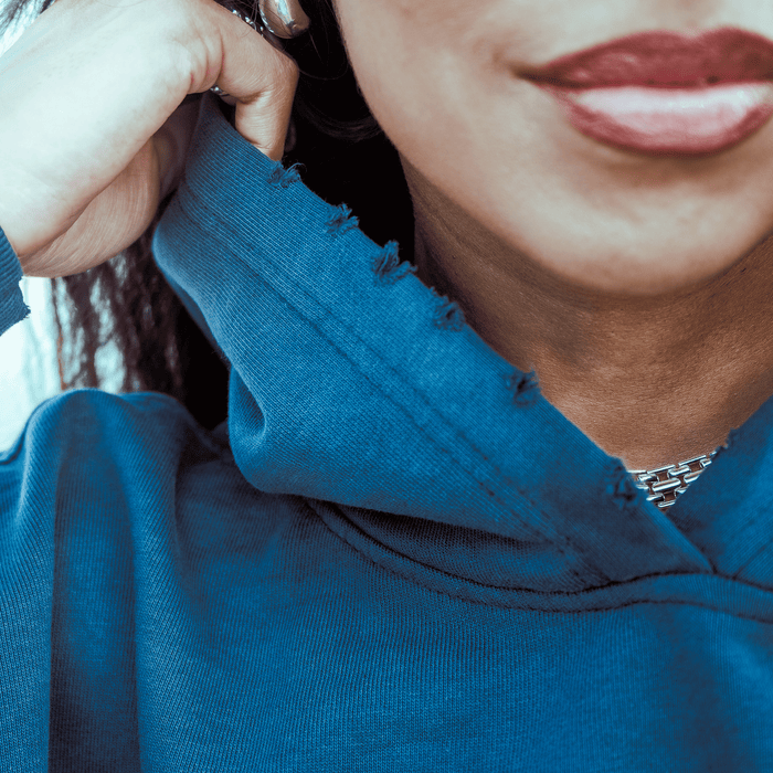 A close-up of a person with medium brown skin wearing bright red lipstick, a silver-link necklace, and the Oaklandish Homage Hoodie with a frayed collar. They adjust the oversized pullover hoodie with one hand.