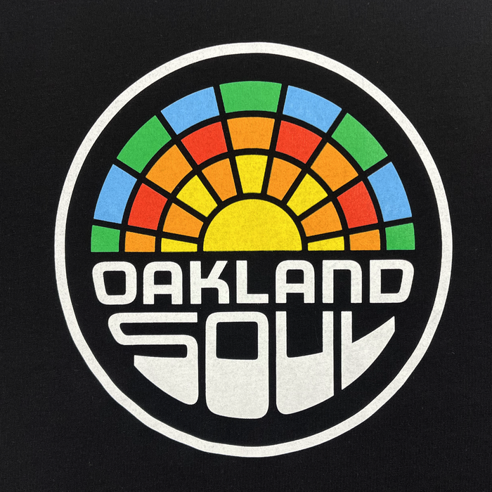 The Infant Oakland Soul Logo One-Piece by Oakland Soul showcases a bold design featuring the words "OAKLAND SOUL" in white, stylized letters at the bottom of a circular logo. Above this text is a vibrant semi-circle resembling a sunset or sun rays with segments in blue, green, yellow, orange, and red. This emblem proudly represents the Oakland Soul women's soccer team in the USL W-League against a sleek black background.
