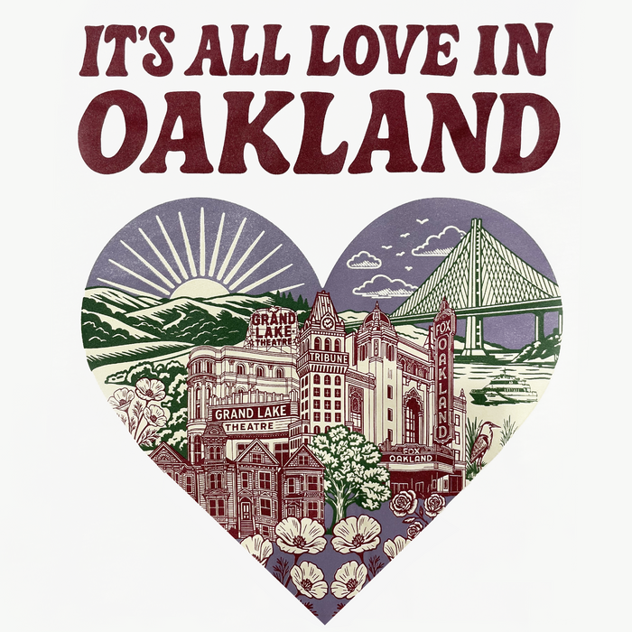 The Oaklandish It's All Love in Oakland Tee features a heart-shaped design with Grand Lake Theatre, Fox Oakland Theatre, Tribune Tower, flowers, and Bay Bridge, plus bold maroon “IT’S ALL LOVE IN THE TOWN” text on a white background.