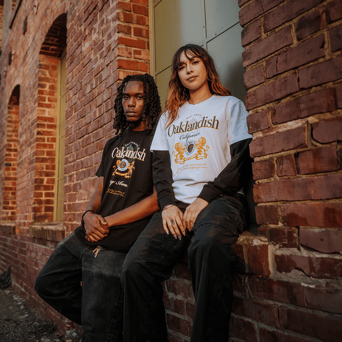 Two people sit on a brick ledge by an old building. The man on the left wears a black Oaklandish Lager Especial Tee, while the woman on the right sports a white classic fit tee over a black long-sleeve. Both face the camera.