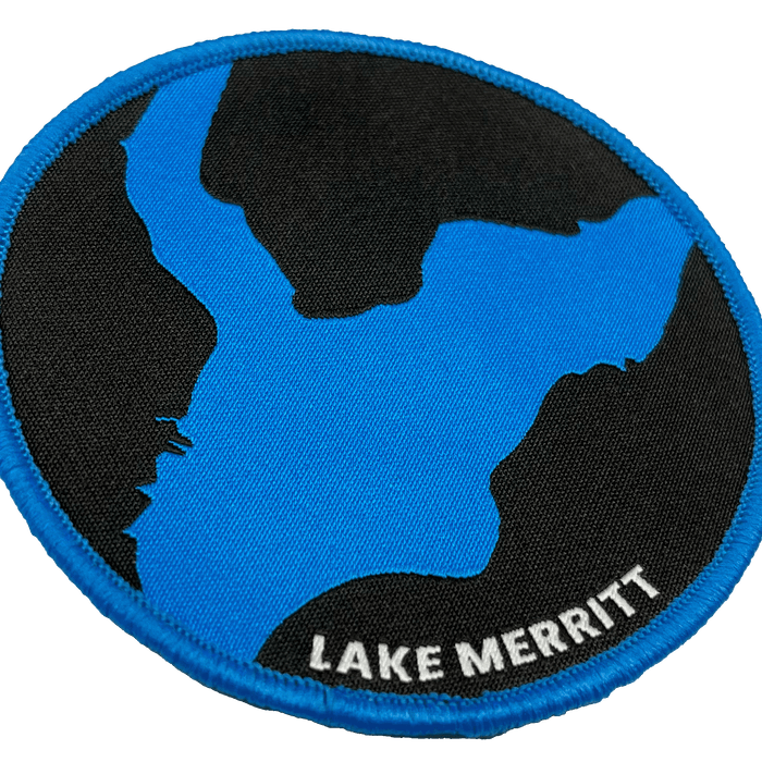 The Oaklandish Lake Merritt Patch features a rounded design with a black background showcasing a blue stylized map of Lake Merritt. The edge is trimmed in blue, and the words "LAKE MERRITT" are elegantly embroidered in white text at the bottom right of this versatile iron-on patch, making it perfect for personalizing your accessories.