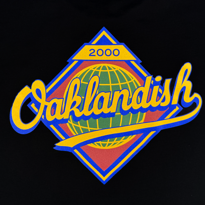 The Leadoff Tee by Other showcases a vibrant design featuring the word “Oaklandish” in yellow cursive lettering across a stylized green globe, all set within a blue and yellow diamond. A yellow rectangle at the top highlights the year “2000.” Printed on a 100% cotton classic fit t-shirt with men’s sizing, this bold graphic pops against the black background.