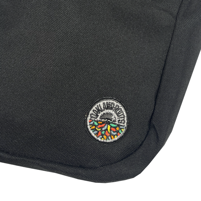 Close-up of the Oakland Roots SC Flight Bag, a black fabric bag featuring a small, round embroidered patch near the bottom corner. The patch showcases a colorful, stylized hedgehog with the text "DON HUSKUTS" around the top. The durable texture and reinforced stitching along the edge make it reminiscent of official gear from an Oakland Roots game.