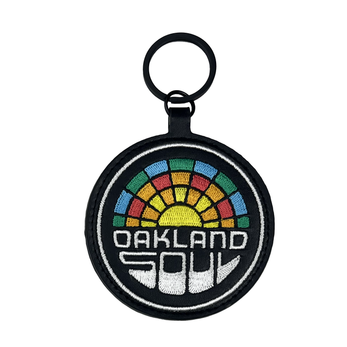The Oakland Soul Logo Keychain, ideal for fans of Oakland's USL W-League women's soccer team, features a round fabric design with a black border. It showcases the words "OAKLAND SOUL" in white block letters at the bottom, and above this text is a colorful geometric design reminiscent of a rainbow mosaic pattern. The keychain is equipped with a black ring for conveniently attaching keys.