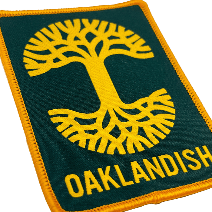 The Oaklandish Rectangle Patch by Oaklandish features a rectangular, embroidered design with a stylized yellow tree with roots at its center against a dark green background. The word "OAKLANDISH" is written in yellow capital letters below the tree. This patch, perfect for accessories, is bordered by yellow stitching and exudes Oakland pride.