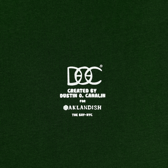 Close-up of white DOC logo Created by Dustin O. Canalin for Oaklandish The Bay-NYC wordmark on a forest green t-shirt.