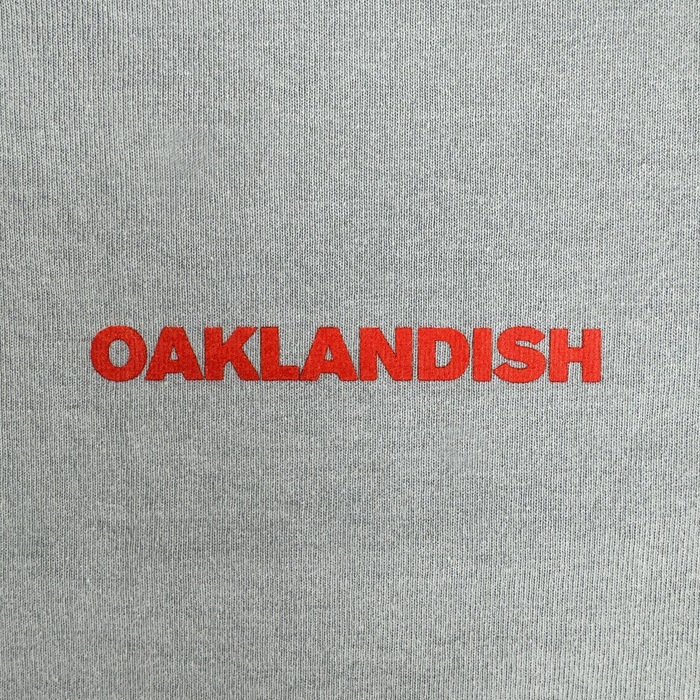 The image features the Oaklandish Tiger Blood Tee: "OAKLANDISH" appears in bold red uppercase letters, centered on a vintage grey, heavyweight cotton shirt with a lightly textured fabric background.