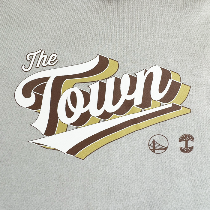 The image features bold, stylized text reading "The Town" in white, brown, and gold on the front of a light gray fabric—likely the Vintage Swash Hoodie by Other. Beneath the text are two small icons: a bridge and a tree. The overall style suggests a sports-inspired or city-themed design.
