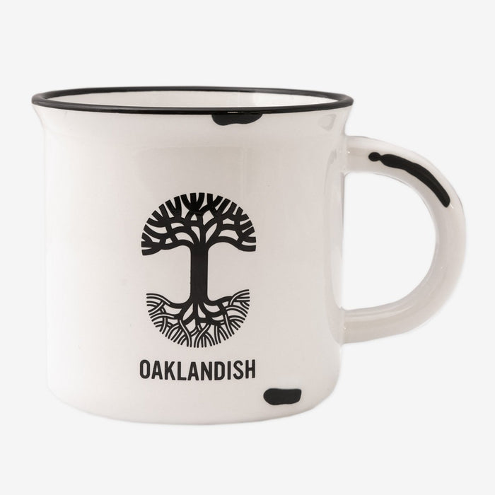 The 15 oz Ventura Mug by Oaklandish is a white ceramic stoneware mug featuring a black rim at the top and a black handle. The mug displays an artistic black tree design with the word "OAKLANDISH" below it, celebrating Oakland city pride. There is a small irregular black spot near the base and handle.