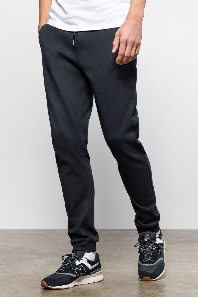 A person wears 686 Mens Everywhere Double Knit Pant in black, paired with a white shirt and black athletic sneakers with white soles, standing on a gray floor against a white background. Only the lower body is shown.