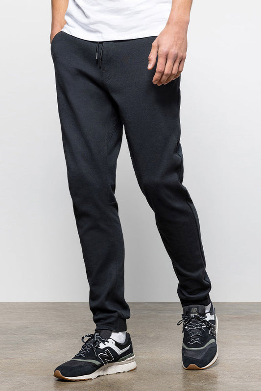 A person wears 686 Mens Everywhere Double Knit Pant in black, paired with a white shirt and black athletic sneakers with white soles, standing on a gray floor against a white background. Only the lower body is shown.