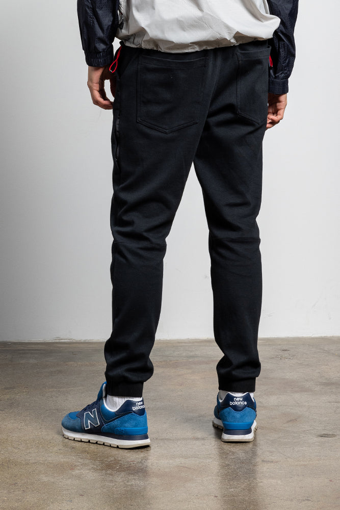 A person stands indoors on concrete, wearing 686 Mens Everywhere Double Knit Pant in black with a light gray jacket, white socks, and blue New Balance sneakers. Photo is from behind, showing only their lower body.