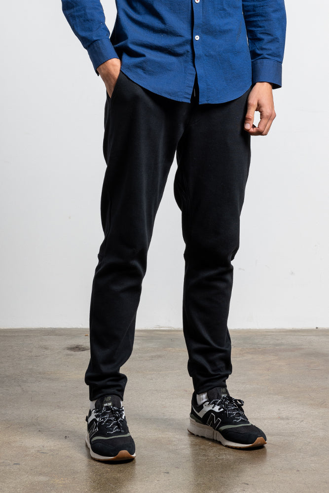 A person stands indoors on a concrete floor, wearing a blue button-up shirt, black 686 Mens Everywhere Double Knit Pant, and black New Balance sneakers. Their hands are in their pockets, and their face is not visible.