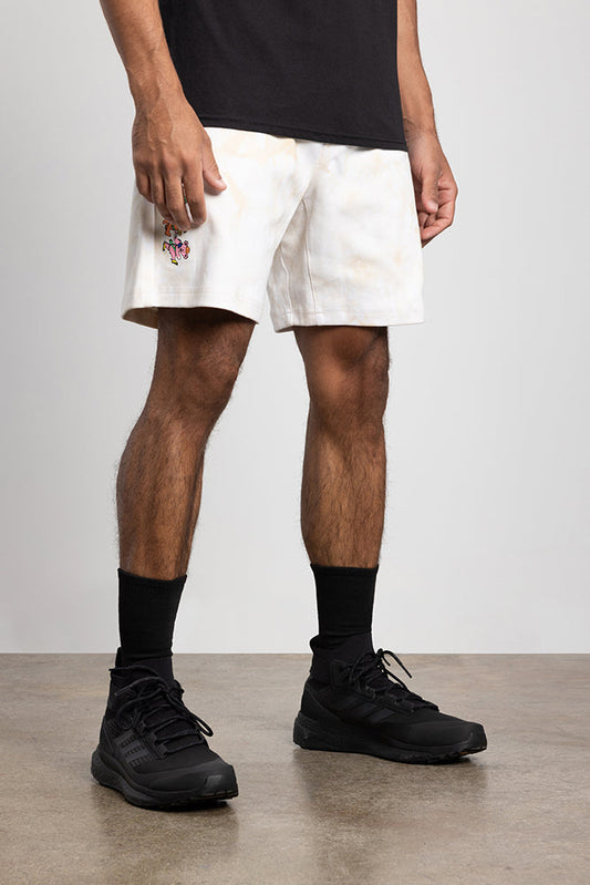 A person stands indoors on a concrete floor wearing a black t-shirt, black socks, black athletic shoes, and 686 Mens Everywhere Double Knit Shorts by 686 in white with a small colorful graphic. The photo is cropped at the shoulders.