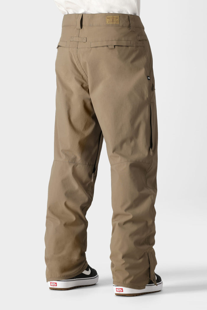 A person wearing 686 Mens Standard Shell Pant by 686 in light brown with secure pockets and black-and-white sneakers stands on a plain white background, showcasing the back view of these loose-fit, affordable shell pants.