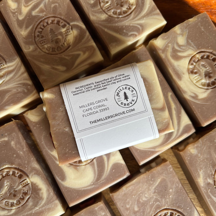 Several rectangular bars of soap with a marbled brown and cream pattern are arranged on a wooden surface. One soap bar has a white paper label wrapped around it with printed text and the Miller's Grove logo.