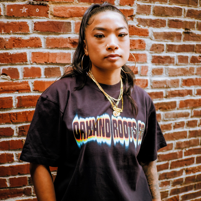 A person with long dark hair in a high ponytail stands against a brick wall, wearing gold hoop earrings, layered gold necklaces, and a black Oakland Roots SC Acid Tee. They look confidently at the camera with a calm expression.