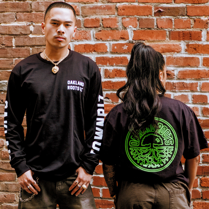 Two people stand by a red brick wall. One faces forward in an Oakland Roots SC Acid Tee, while the other displays the neon green Oakland Roots tree logo on the back of their Oaklandish tee.