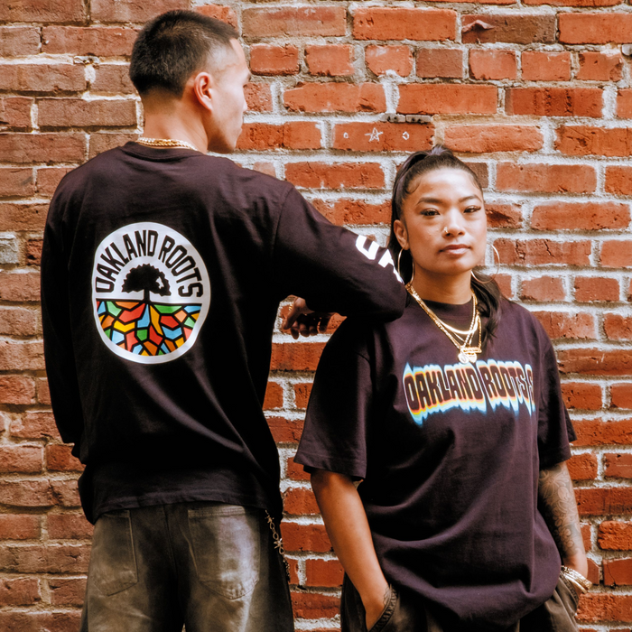 Two people stand by a brick wall. One, facing away, wears the Oakland Roots SC Acid Tee with a colorful tree logo. The other faces forward in another bold Oakland Roots top. Both have gold jewelry and dark casual pants.