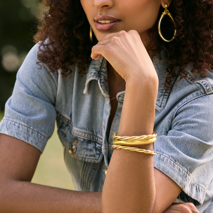 A close-up lifestyle shot of a woman wearing a stack of three gold bangle bracelets on her wrist, paired with a light blue denim shirt and gold hoop earrings.