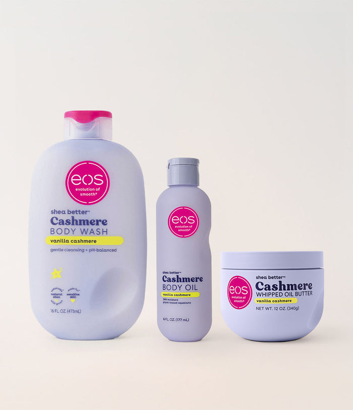Three EOS Shea Better body care products in a clean studio setup: a large Cashmere Body Wash bottle with a pink cap, a smaller Cashmere Body Oil bottle with a gray cap, and a Cashmere Whipped Oil Butter jar with a white lid, all on a light beige background.
