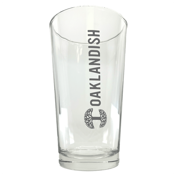 The Banner Pint Glass from Oaklandish is a clear drinking vessel with the word "OAKLANDISH" vertically inscribed in bold, black letters. Underneath this text, you'll find the brand's dark grey logo, a detailed circular tree design. The drinkware has a smooth, slightly tapered shape and features a thick base.