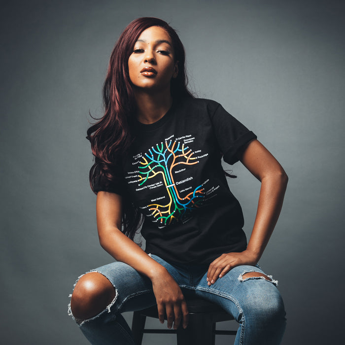 A woman with long, wavy auburn hair sits confidently on a stool against a gray background. She's wearing Oaklandish's BART Tree Tee, a classic fit black T-shirt featuring a colorful tree design with various words on its branches, paired with ripped blue jeans. Her expression is self-assured, with her head slightly tilted.