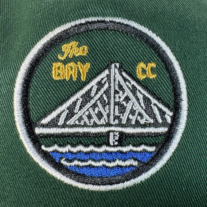 The Bay Area 5 Panel By DOC from Oaklandish features an embroidered white bridge over blue water on dark green fabric, bordered in black and white with "The BAY CC" in yellow—perfect for your favorite 5-panel hat.