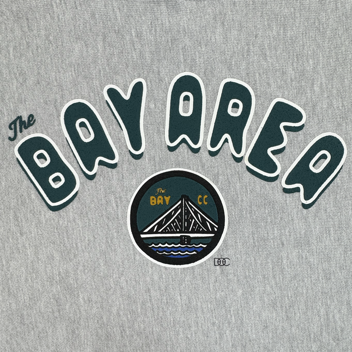 Sure! Here’s the sentence rewritten using your product data: A gray background with bold, rounded green and white text spells out “The Bay Area.” Beneath it, a circular emblem features a bridge over water with “The Bay” and “CC” in yellow lettering, symbolizing the San Francisco Bay Area. This design appears on the heavyweight fleece pullover Bay Area CC Hoodie By DOC from Other’s The Bay Collection.