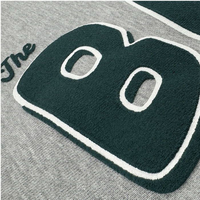 A textured green applique of the number “80” with white borders is sewn onto heavyweight heather gray fleece fabric. The word “The” appears in cursive to the left of the numbers, also in green, referencing The Bay Collection. This soft and fuzzy surface makes the Bay Area CC Hoodie By DOC by Other perfect for a cozy pullover hoodie.