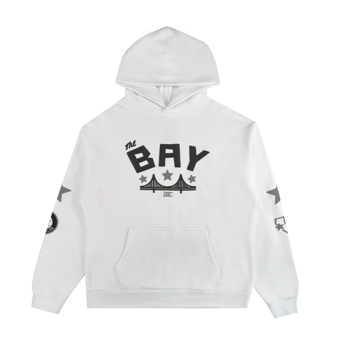 The Bay Bridge Hoodie By DOC V2 by Oaklandish is a white pullover with a front pocket, "THE BAY" in bold black letters above a bridge graphic, black stars on the sleeves, and a black-and-white patch on the right sleeve.