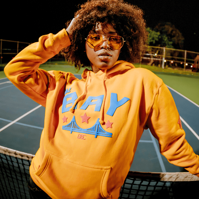 Wearing the Bay Bridge Hoodie by DOC from Other—a bold orange piece featuring “E BAY” text, blue graphics, and red stars—a person poses confidently by a tennis net at night, large amber-tinted sunglasses accenting their look and one hand resting on their head. With the tennis courts and trees as a backdrop, this eye-catching scene channels the essence of an art project rooted in Bay Area street style.