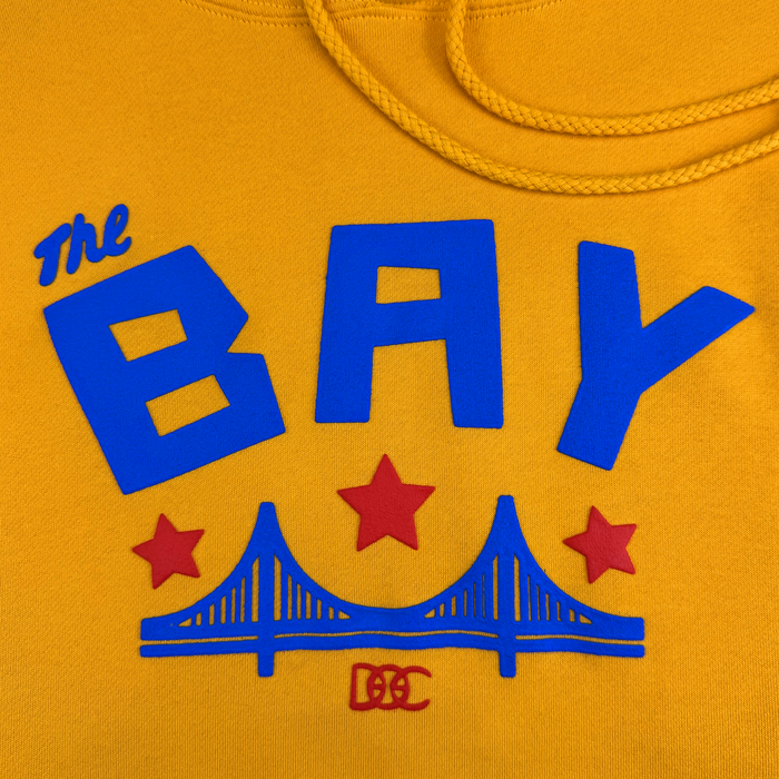 Introducing the Bay Bridge Hoodie by DOC from Other: This vibrant yellow designer hoodie features “The BAY” in bold blue lettering, highlighted by three striking red stars and a stylized blue Golden Gate Bridge graphic. A small red “DC” logo is displayed beneath the art, while a matching yellow drawstring completes the look—a perfect tribute to the Bay Area and an ideal centerpiece for any creative project.