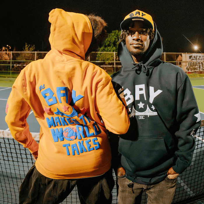 Two people stand by a tennis court net at night. One faces forward, wearing a dark hoodie and yellow “BAY” cap. The other faces away in the Bay Bridge Hoodie by DOC from Other—a designer orange hoodie featuring colorful “The BAY Makes It, The World Takes” text and globe graphics—showcasing Bay Area creativity as part of an art project.