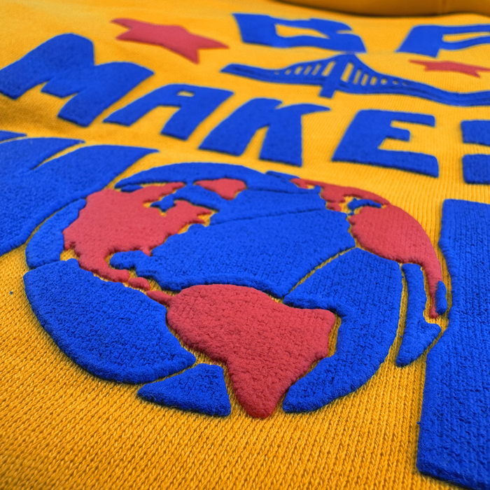 Close-up of yellow fabric embroidered with blue and red details. The design features a globe with blue oceans and red continents, along with bold blue text reading “MAKE E”. Above the text are two red stars and a stylized bridge inspired by the Bay Area, giving this Bay Bridge Hoodie by DOC from Other an artful, project-inspired aesthetic.