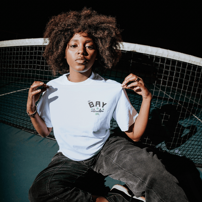 A woman with an afro hairstyle kneels on a tennis court at night, pulling at the front of her white “Bay Bridge Tee by DOC” from Other. She pairs the heavyweight cotton t-shirt with dark jeans and gazes confidently at the camera, the tennis net and court lines forming a compelling backdrop—an expressive scene reminiscent of an art project inspired by the Bay Area.