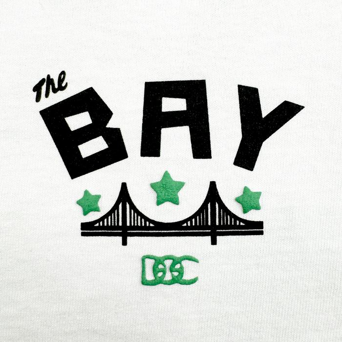 The Bay Bridge Tee by DOC from Other is a white heavyweight cotton t-shirt featuring bold black “The BAY” text above a minimalist black bridge graphic, inspired by the Bay Area. Three green stars sit above the bridge, with a green stylized “DC” logo below. The clean design draws inspiration from art projects and uses minimal colors.