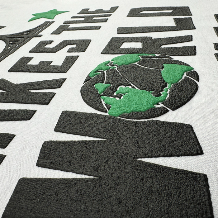 Close-up of textured black and green embroidered lettering on heavyweight cotton fabric from the Bay Bridge Tee by DOC (brand: Other). The design features a stylized green globe depicting Asia, Australia, and part of Africa, alongside bold partial text reading “KES THE WO,” with a prominent green star above—evoking a creative art project inspired by Bay Area style.