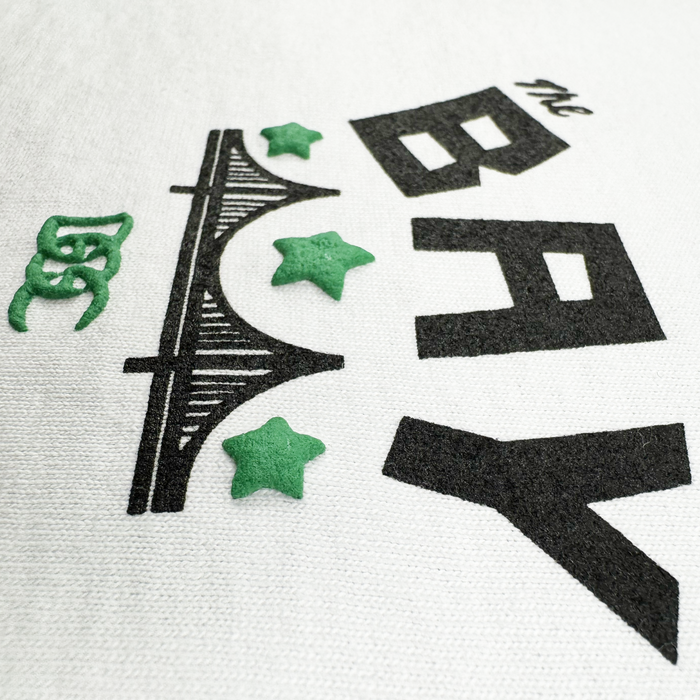 A close-up of the Bay Bridge Tee by DOC from Other, a heavyweight cotton t-shirt, featuring “The BAY” in bold black text above a black stylized bridge and three green 3D stars. Set against a white background that captures the spirit of the Bay Area, the design also includes a small decorative green symbol on the left side near the bridge.
