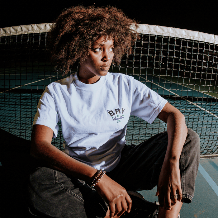 A woman with curly hair, dressed in the Bay Bridge Tee by DOC from Other and dark jeans, sits cross-legged on a tennis court at night. She gazes to her left, resting one arm on her knee, while the tennis net and court lines appear in the background—capturing an artful Bay Area moment.