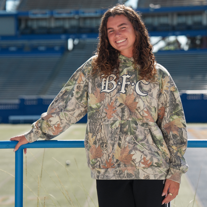 A young woman with curly brown hair smiles outside in the Bay FC Camo Hoodie by Bay FC, featuring "B.F.C" on the front. She leans on a blue railing near empty stadium bleachers and a sports field in bright sunlight. Men's sizing available.