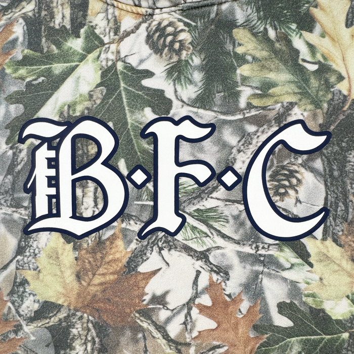 The image features the letters "B.F.C" in a bold, black-outlined white font over fall leaves, pinecones, and wood camo in green, brown, and tan—ideal for the Bay FC Camo Hoodie by Bay FC.
